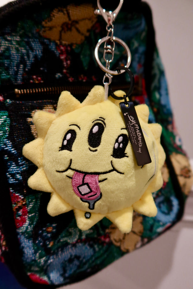 Plush sun-shaped keychain with a tongue sticking out on a colorful bag.