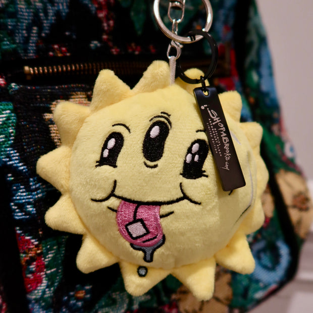 Plush sun-shaped keychain with a tongue sticking out on a colorful bag.